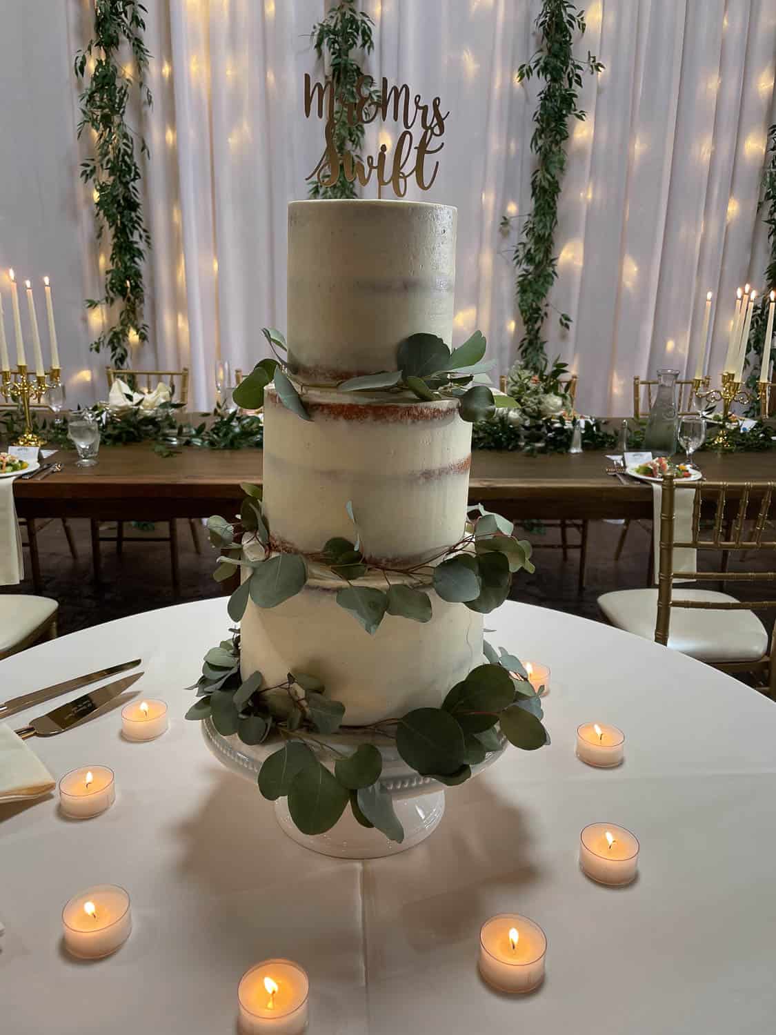 Three-tiered semi-naked wedding cake decorated with eucalyptus leaves and a “Mr and Mrs Swift” topper, surrounded by lit tea candles on a round table.