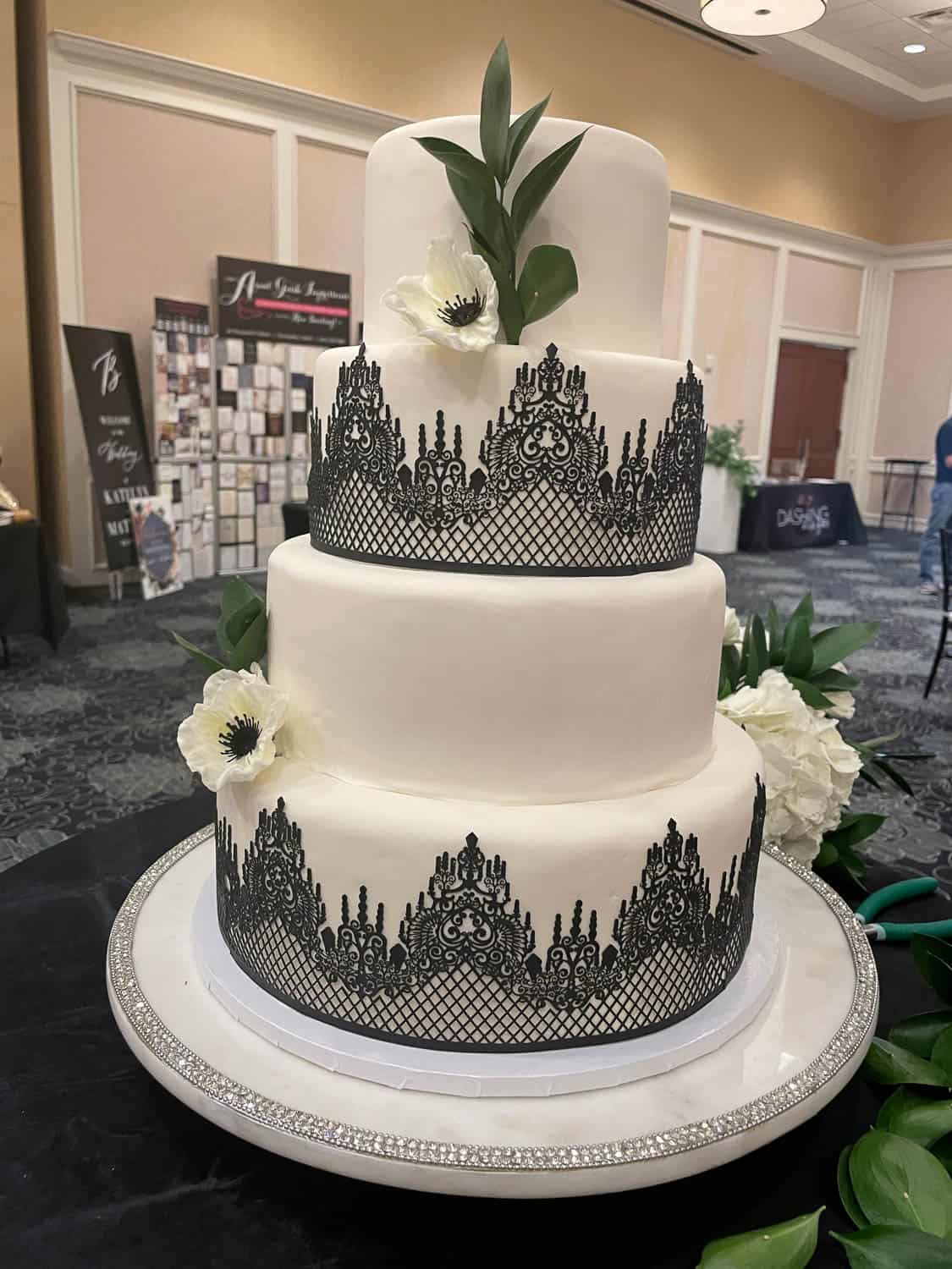 Three-tier white cake decorated with black lace-like edible accents, white flowers, and green leaves, displayed on a round tray with a jeweled edge.