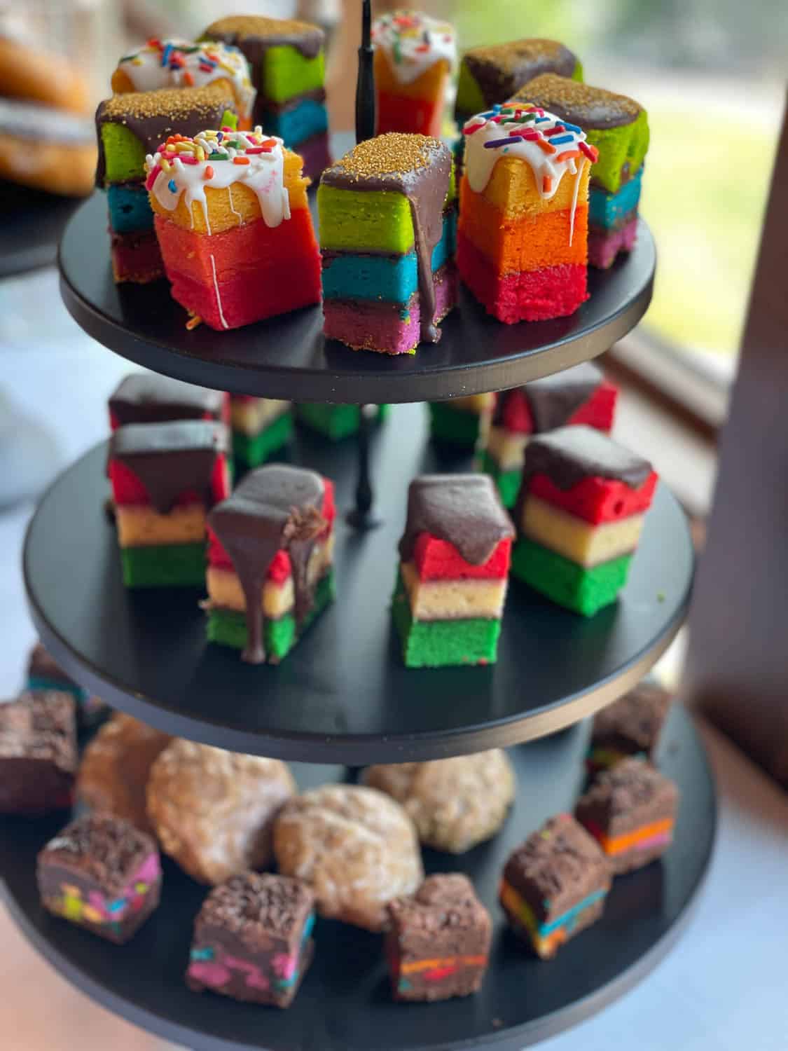 A three-tiered tray displaying colorful layered rainbow cakes, chocolate-covered layered squares, and round cookies with sprinkled toppings.