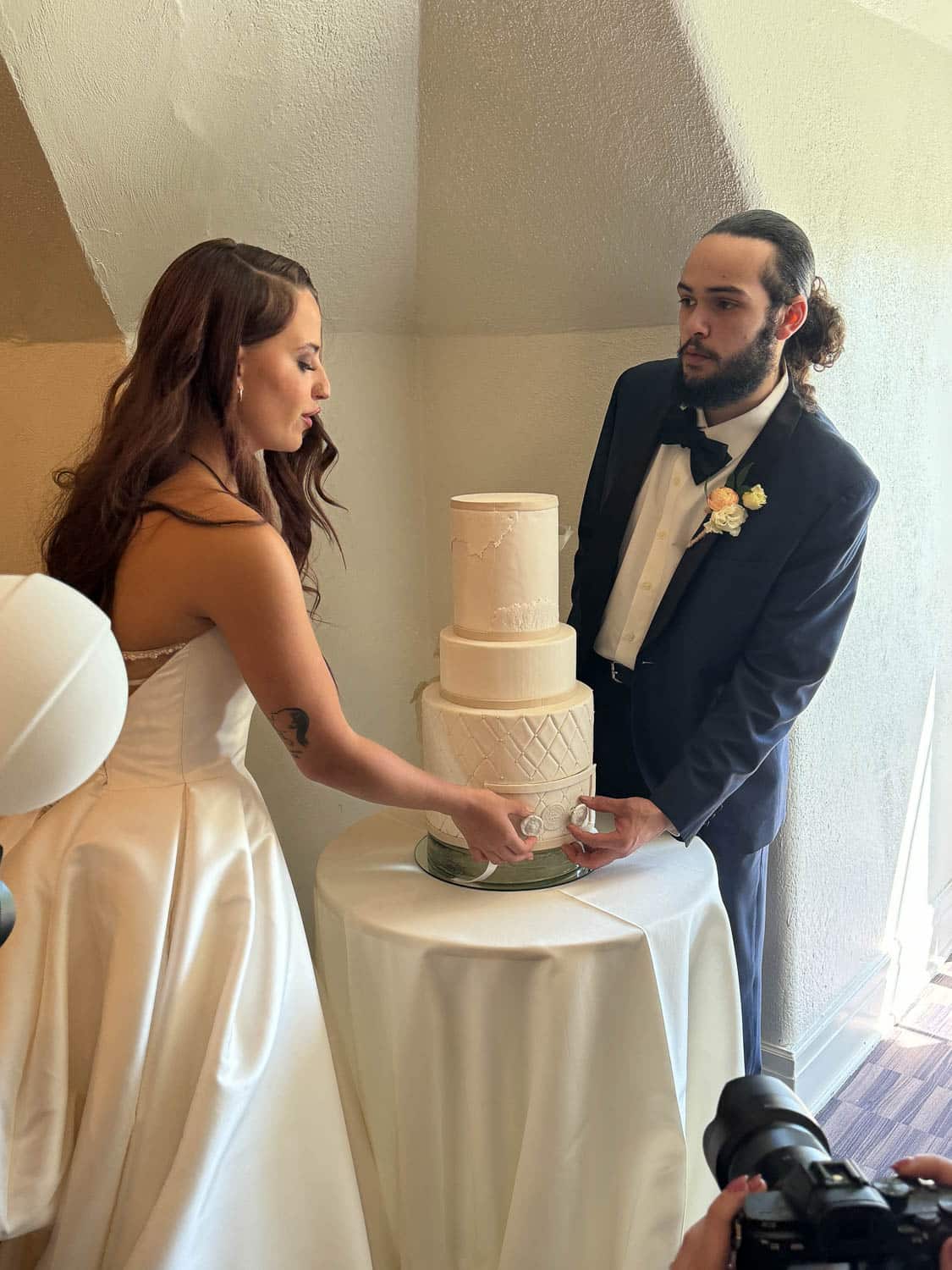 A bride and groom stand together, with a three-tier white wedding cake on a small round table, with a photographer capturing the moment.
