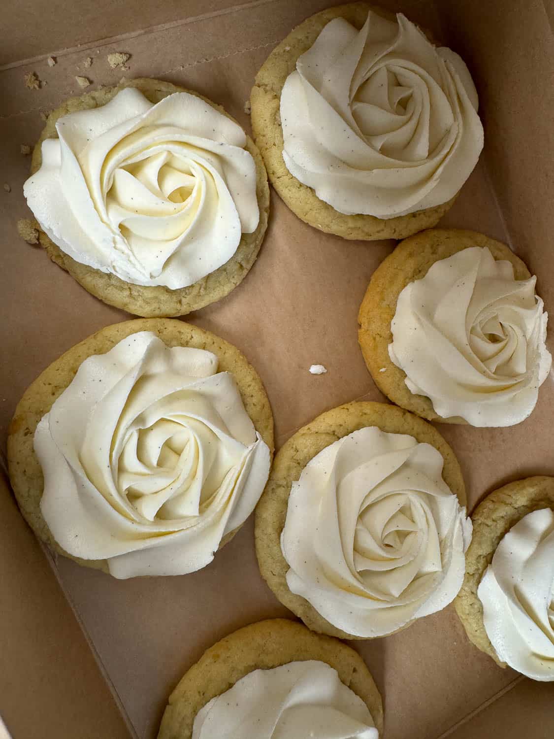 Seven sugar cookies with white rose-shaped frosting arranged inside a brown cardboard box.