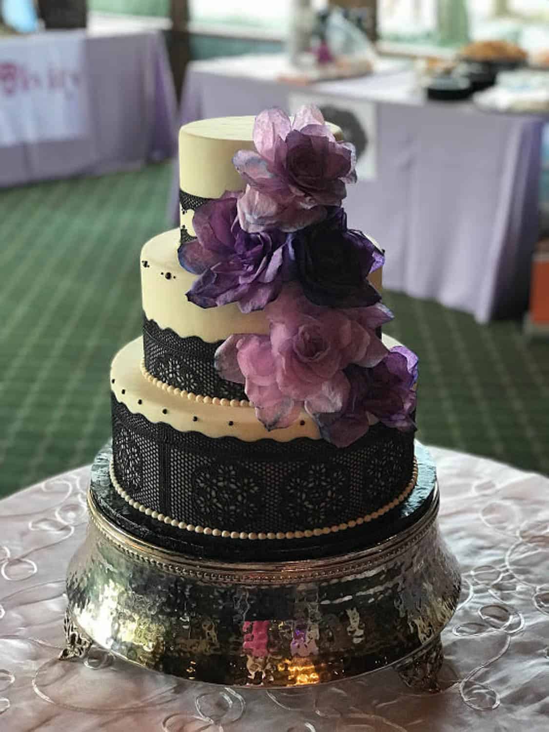 A three-tiered cake decorated with black lace, pearl borders, and large purple flowers, displayed on a metallic stand.