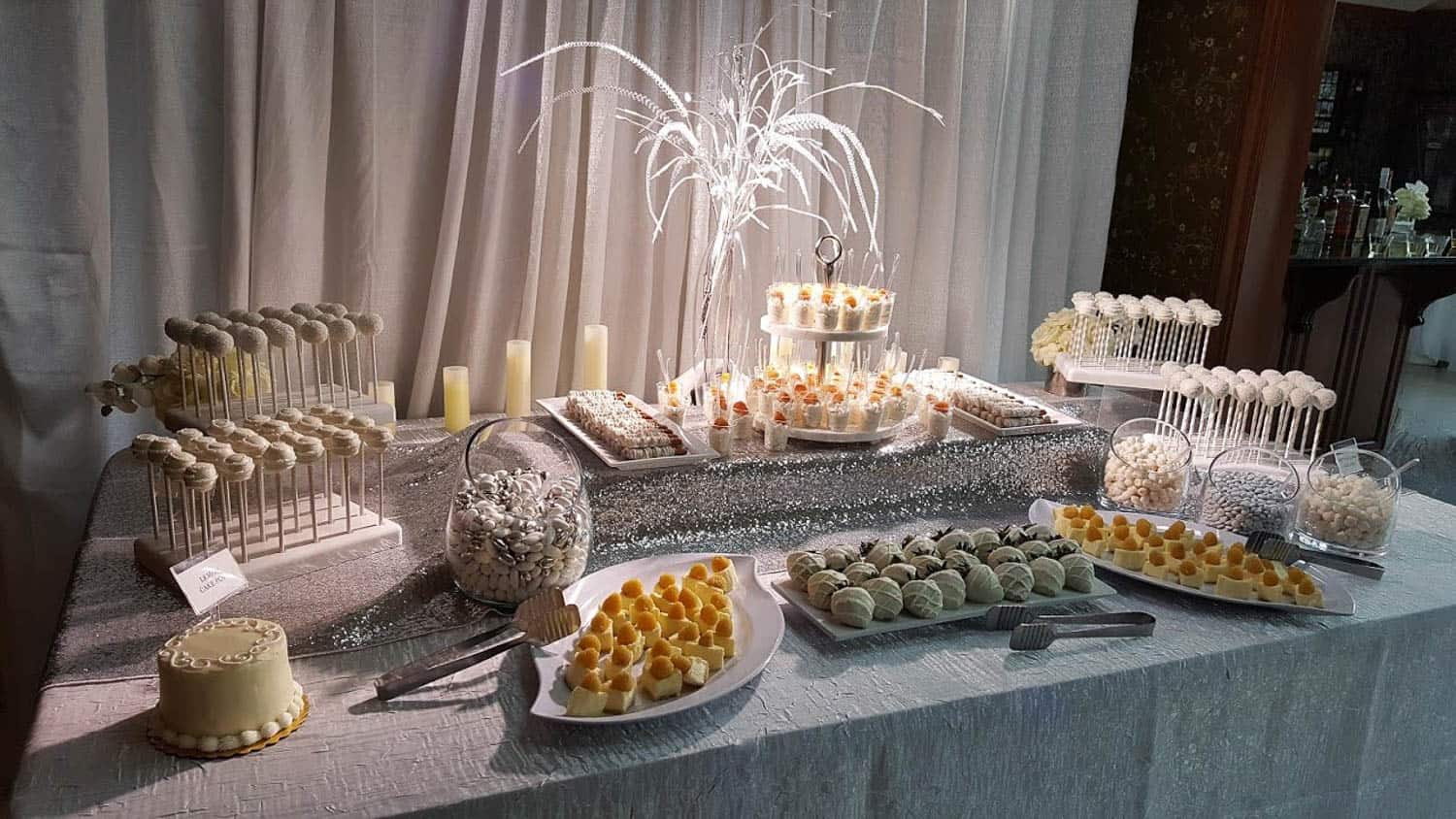 Dessert table with cake pops, small cakes, truffles, and candies on white plates and stands, accented by a decorative centerpiece and candles.