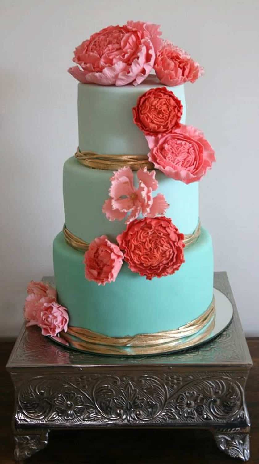 Three-tiered cake with light blue fondant, gold ribbon accents, and large pink and red decorative flowers, displayed on an ornate silver cake stand.