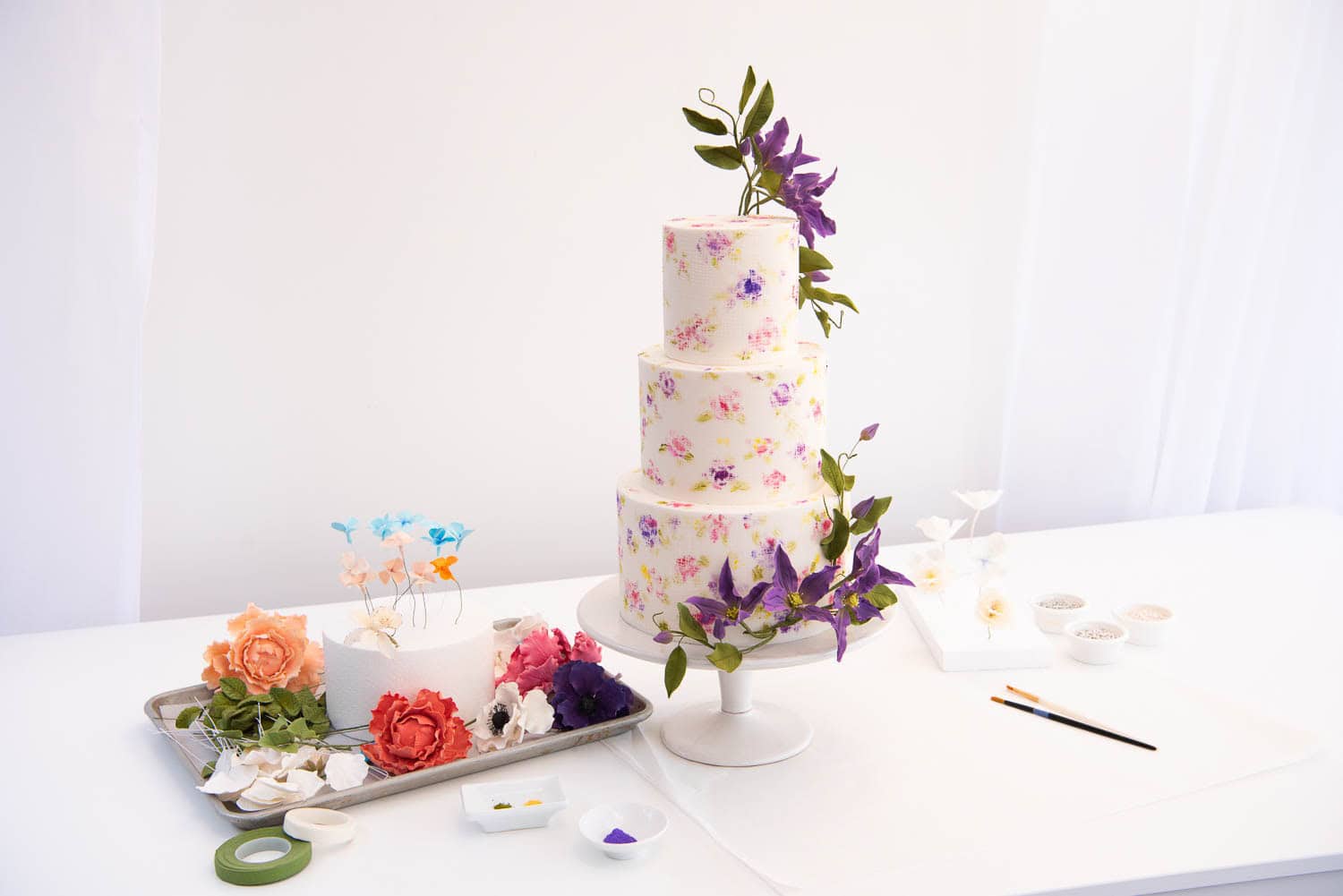 A three-tiered white cake with purple floral decorations on a stand, surrounded by sugar flowers, art supplies, and a paintbrush on a white table.