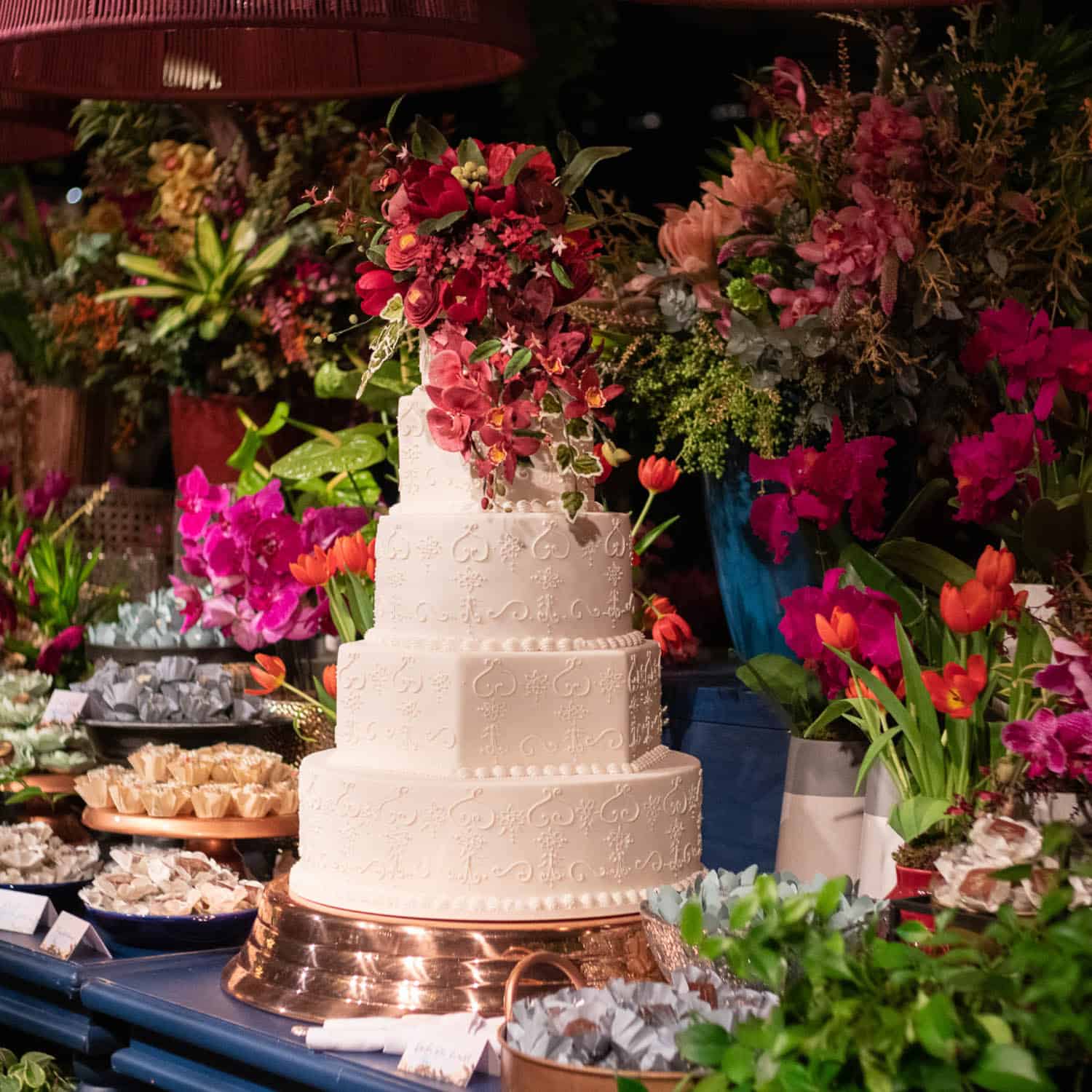 A four-tiered white wedding cake decorated with cascading flowers is displayed on a copper stand, surrounded by assorted colorful flower arrangements and sweets.