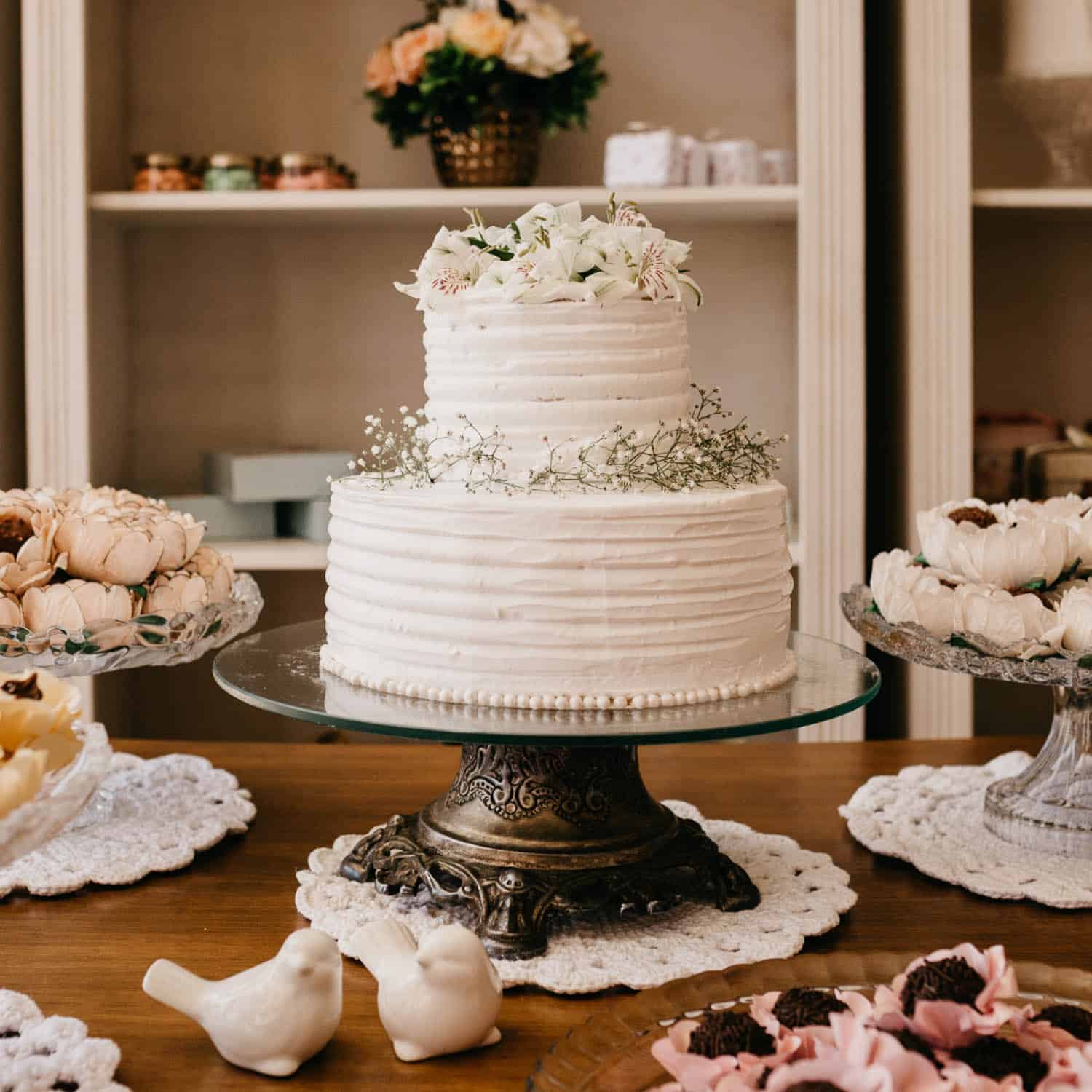 Two-tiered white frosted cake with floral decorations on a glass stand, surrounded by assorted desserts and white ceramic bird figurines on a wooden table.