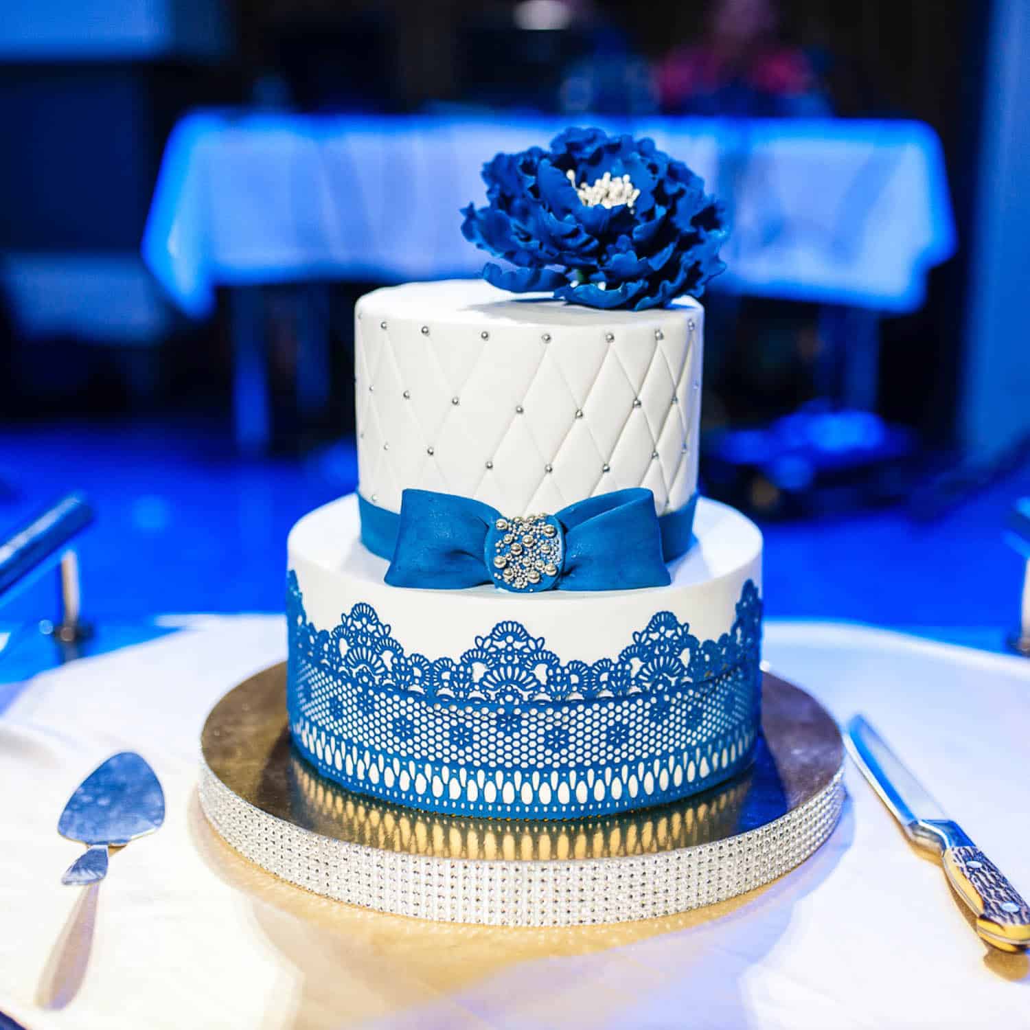 Two-tiered white cake decorated with blue lace, a blue bow with a jeweled brooch, and a large blue flower on top, placed on a round table with cutlery.