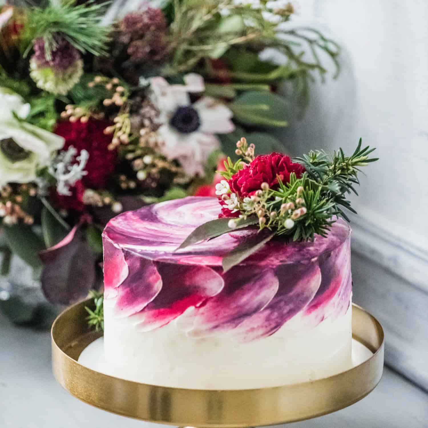 A round cake with white and maroon swirled frosting, topped with rosemary and red flowers, sits on a gold stand with a floral arrangement in the background.