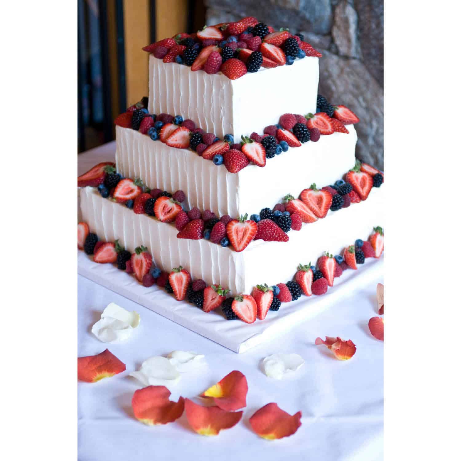 Three-tiered square white cake topped and decorated with assorted fresh berries, displayed on a white tablecloth with scattered rose petals.