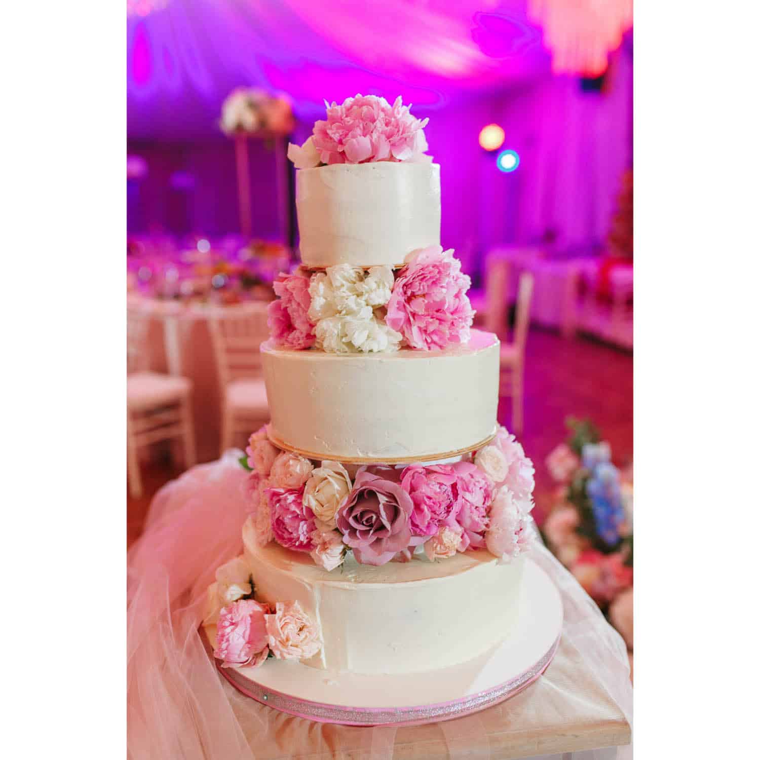 Four-tier white cake decorated with pink and white flowers sits on a table at an event with purple ambient lighting.