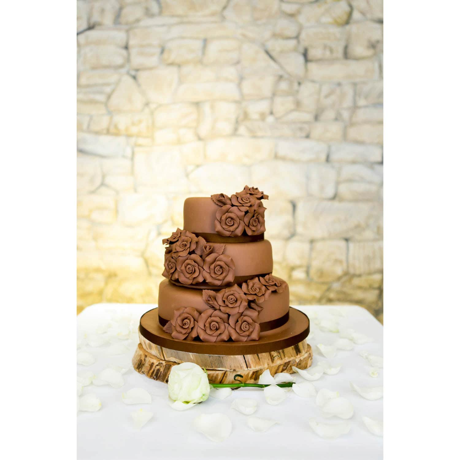 Three-tiered chocolate cake decorated with chocolate roses, displayed on a wooden stand surrounded by white rose petals, against a stone wall background.