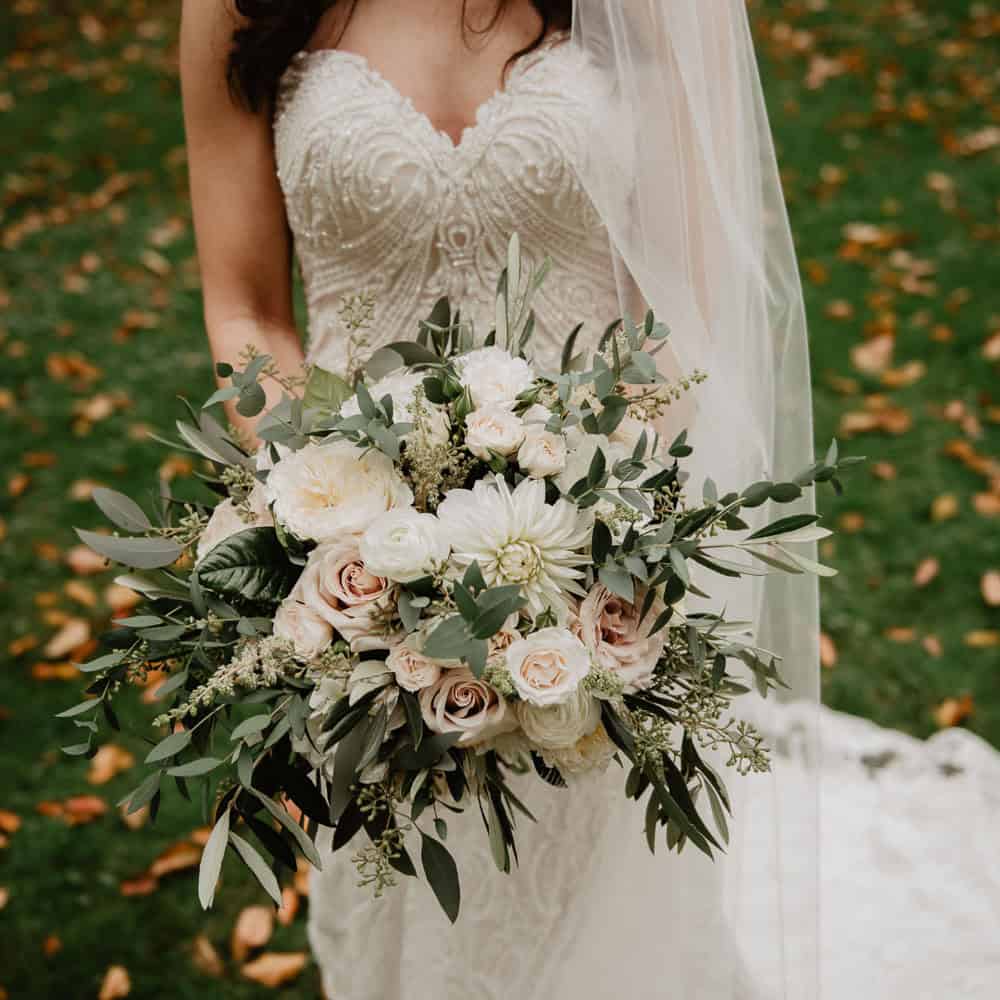 Bride in a white lace dress holds a large bouquet of white and pale pink flowers with greenery. Fallen leaves and green grass are visible in the background.