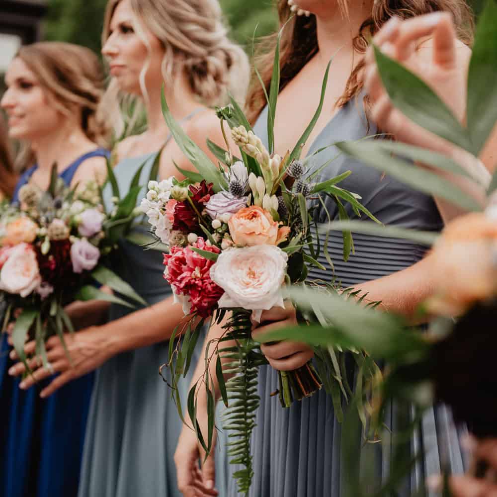 Four women in bridesmaid dresses holding colorful flower bouquets, standing side by side at an outdoor event.