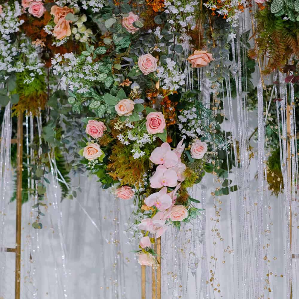 A floral arrangement with pink roses, white flowers, greenery, and hanging strands of clear beads against a light backdrop.