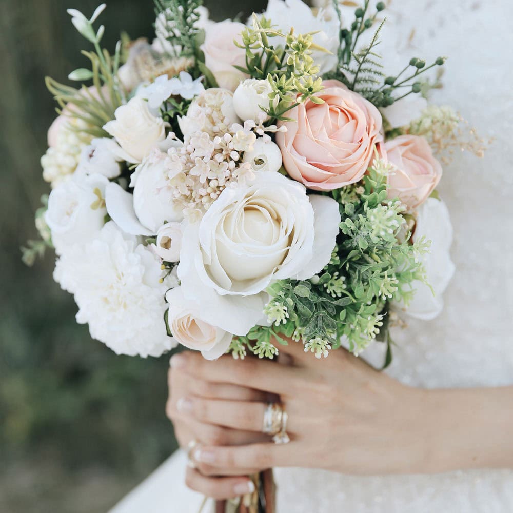 A woman in a white dress holds a bouquet of pastel roses, white flowers, and greenery, with wedding rings visible on their fingers.
