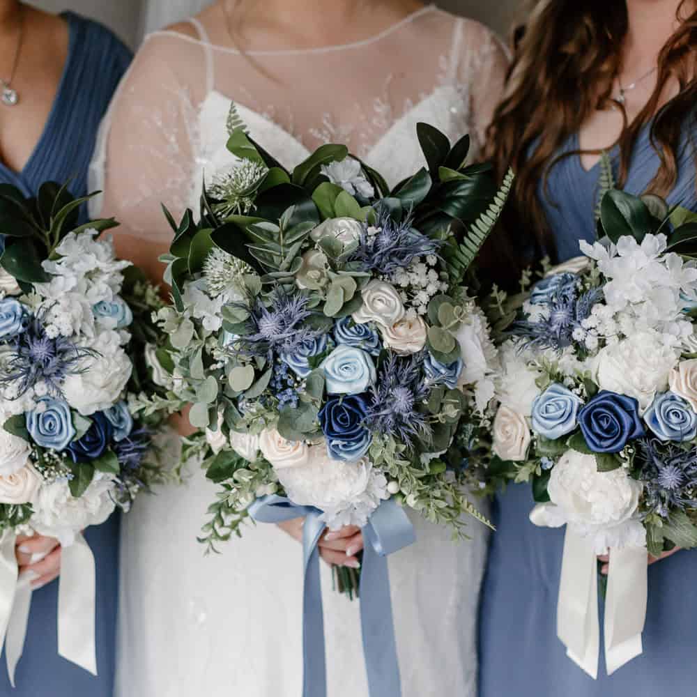 Two women in blue and a bride in white, hold bouquets with blue, white, and cream flowers, featuring greenery and ribbon accents.
