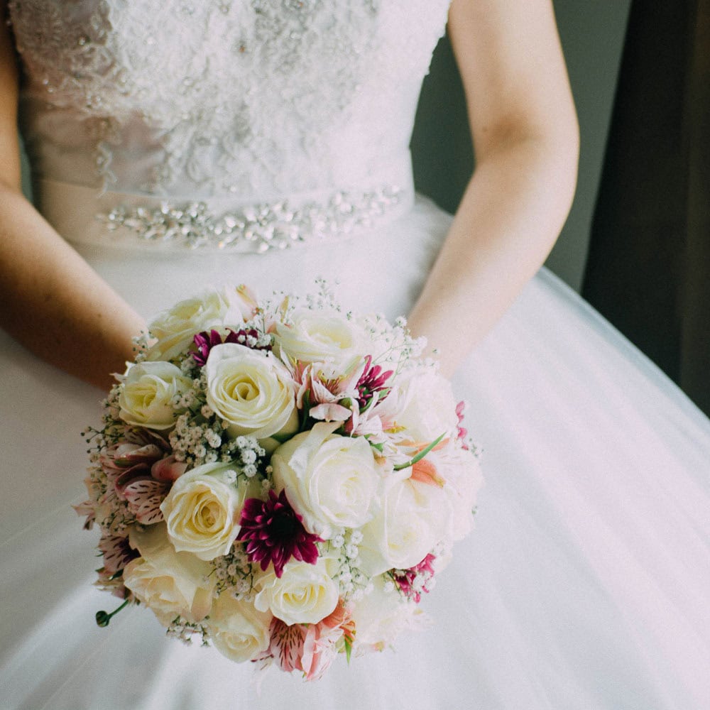 A bride in a white lace wedding dress holds a bouquet of white roses and pink flowers, photographed from the waist down.