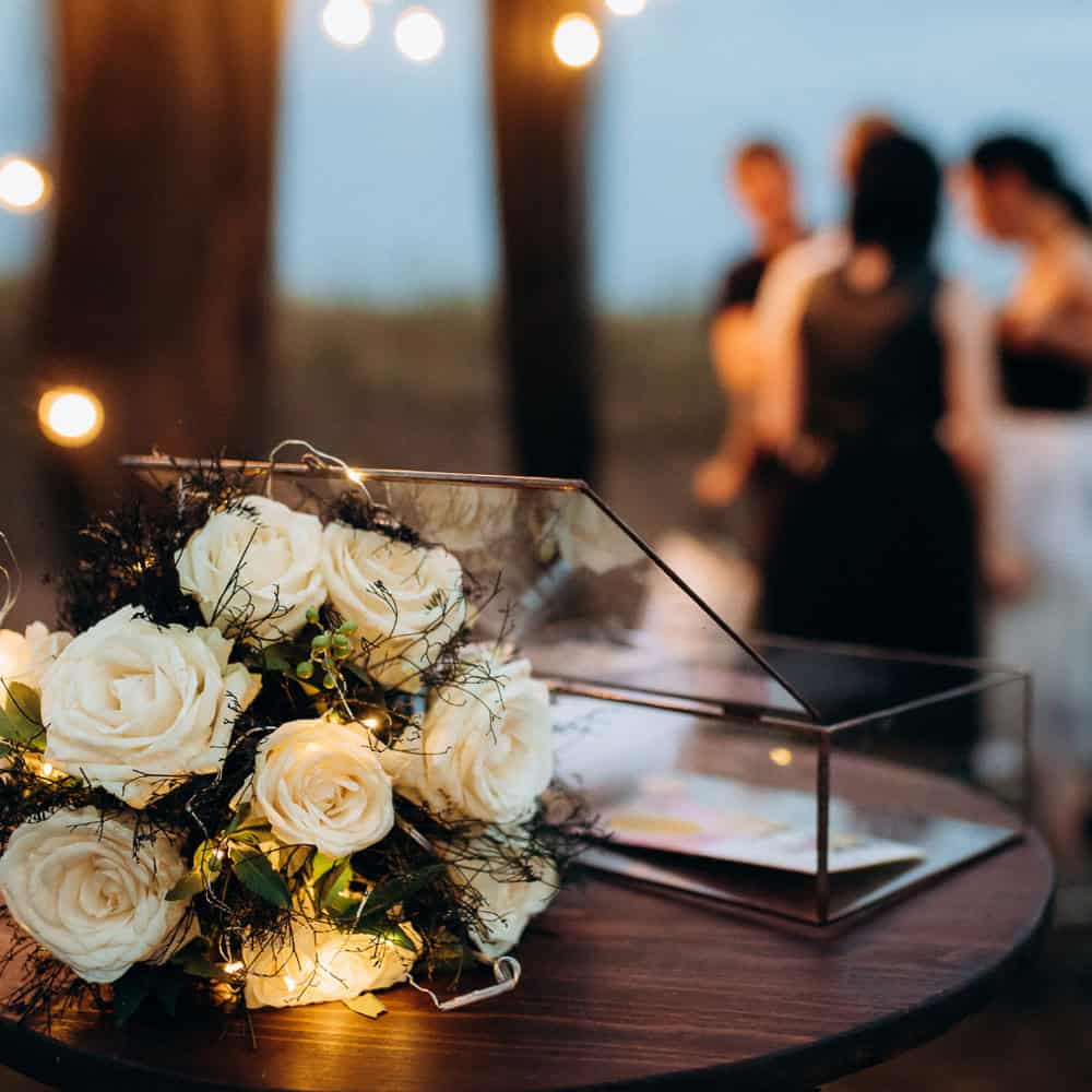 A bouquet of white roses rests on a table with a glass box, while four people stand blurred in the background under string lights.