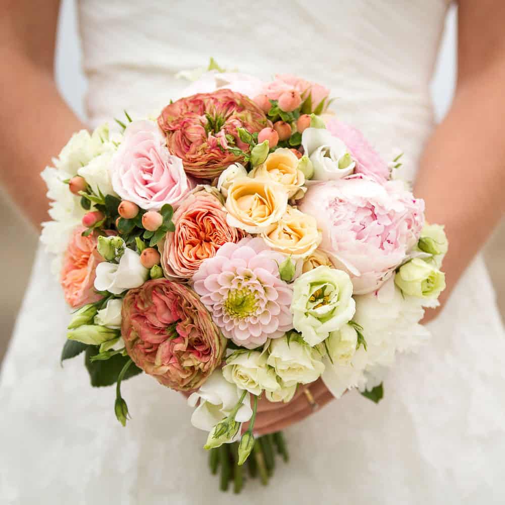 A woman in a white dress holds a bouquet of assorted flowers, including roses, dahlias, and peonies, in soft pink, peach, and cream tones.