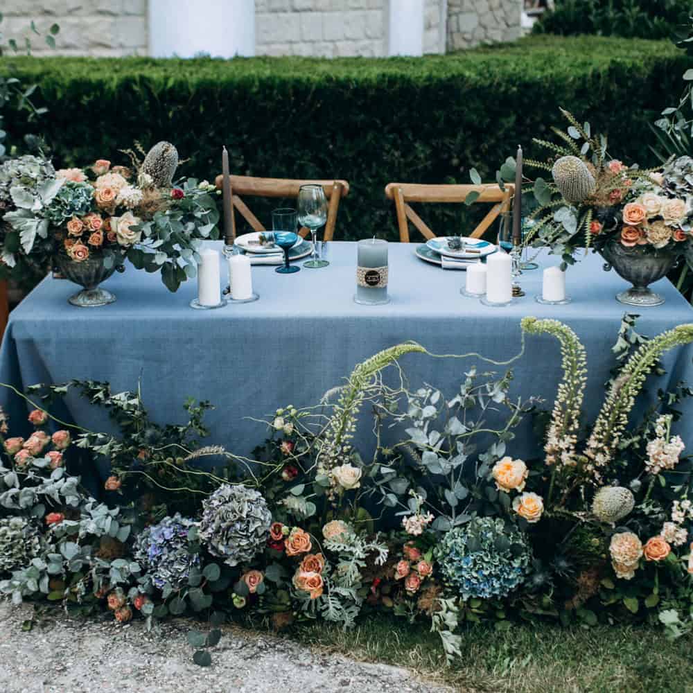 A decorated outdoor table with a blue tablecloth, floral centerpieces, candles, plates, glasses, and an arrangement of flowers and greenery along the front edge.