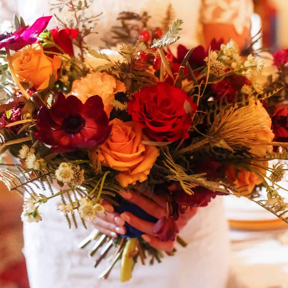 A woman in a white outfit holds a large bouquet of red, yellow, and orange flowers with greenery.