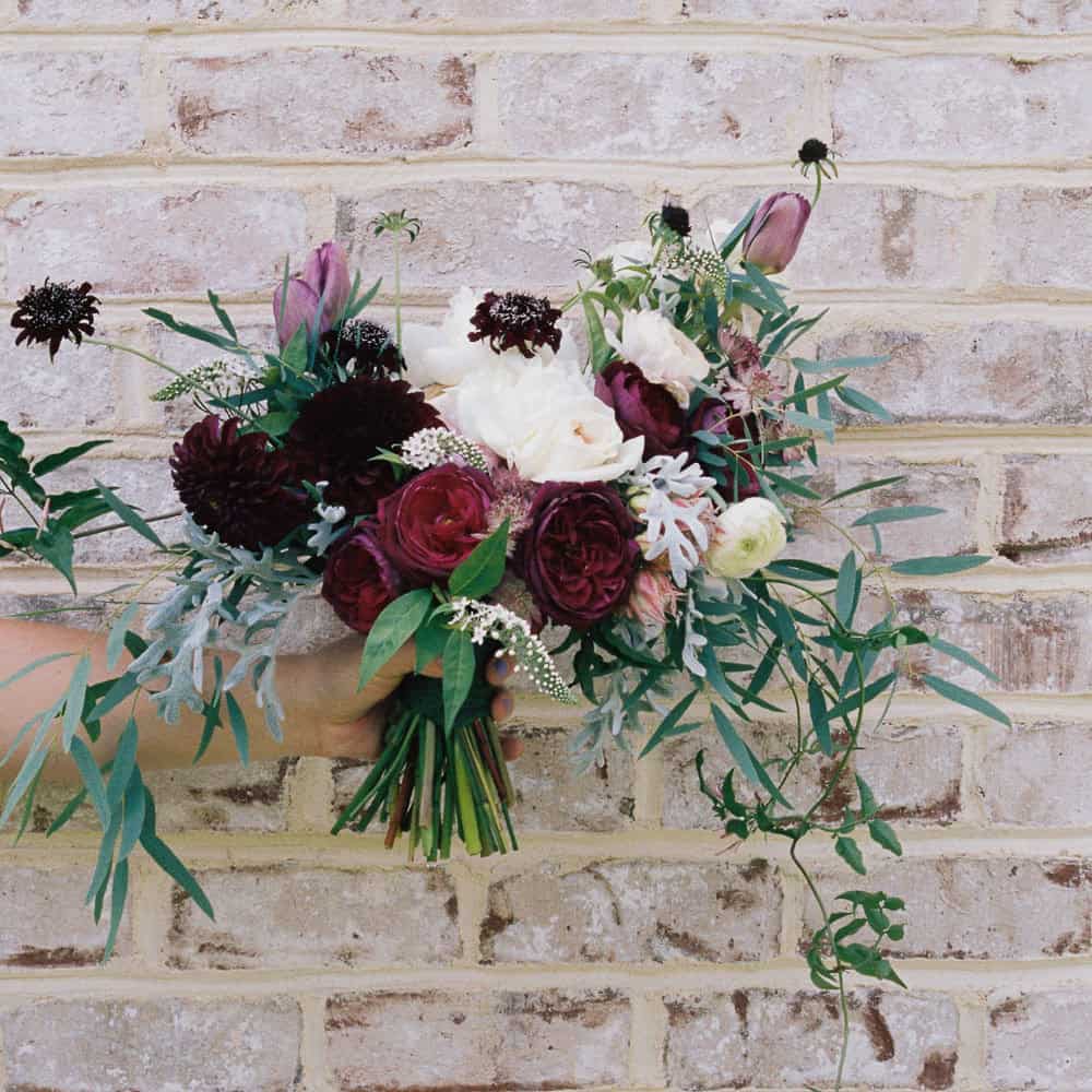 A hand holding a bouquet of flowers with burgundy, white, and pink blooms, accented by green foliage, in front of a light brick wall.