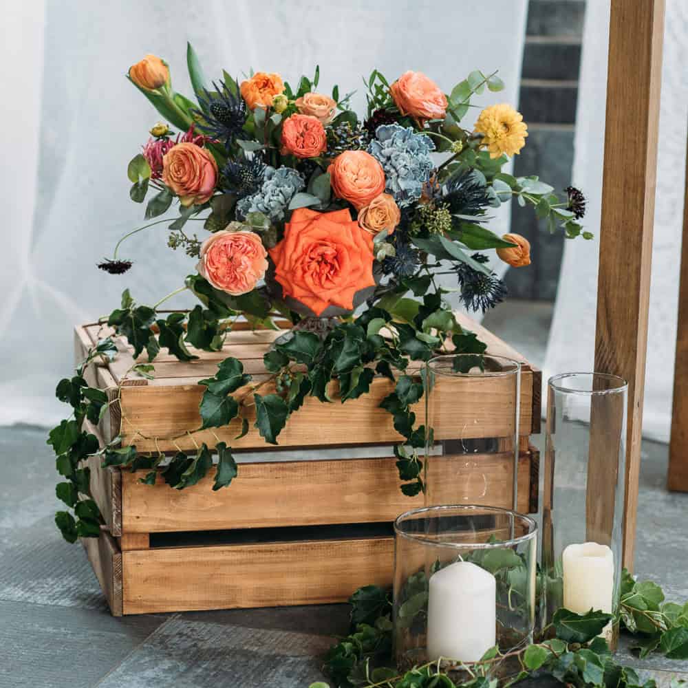 A wooden crate holds a colorful flower arrangement with orange, yellow, and blue blooms, green foliage, and trailing ivy, next to glass candles on the floor.