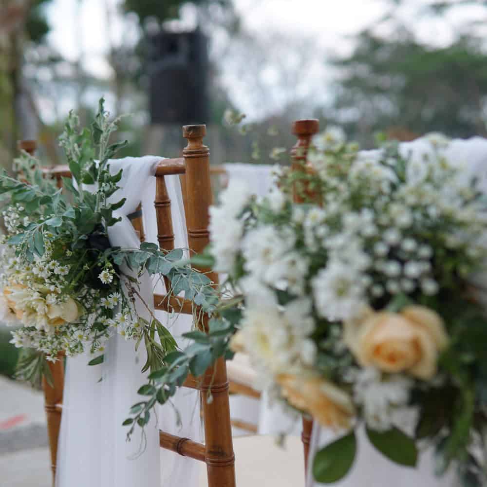 Close-up of wooden chairs decorated with white fabric, greenery, and pale yellow flowers, set up outdoors.