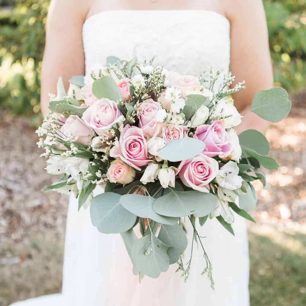 A bride in a strapless white dress holds a bouquet of pink roses, white flowers, and greenery outdoors.