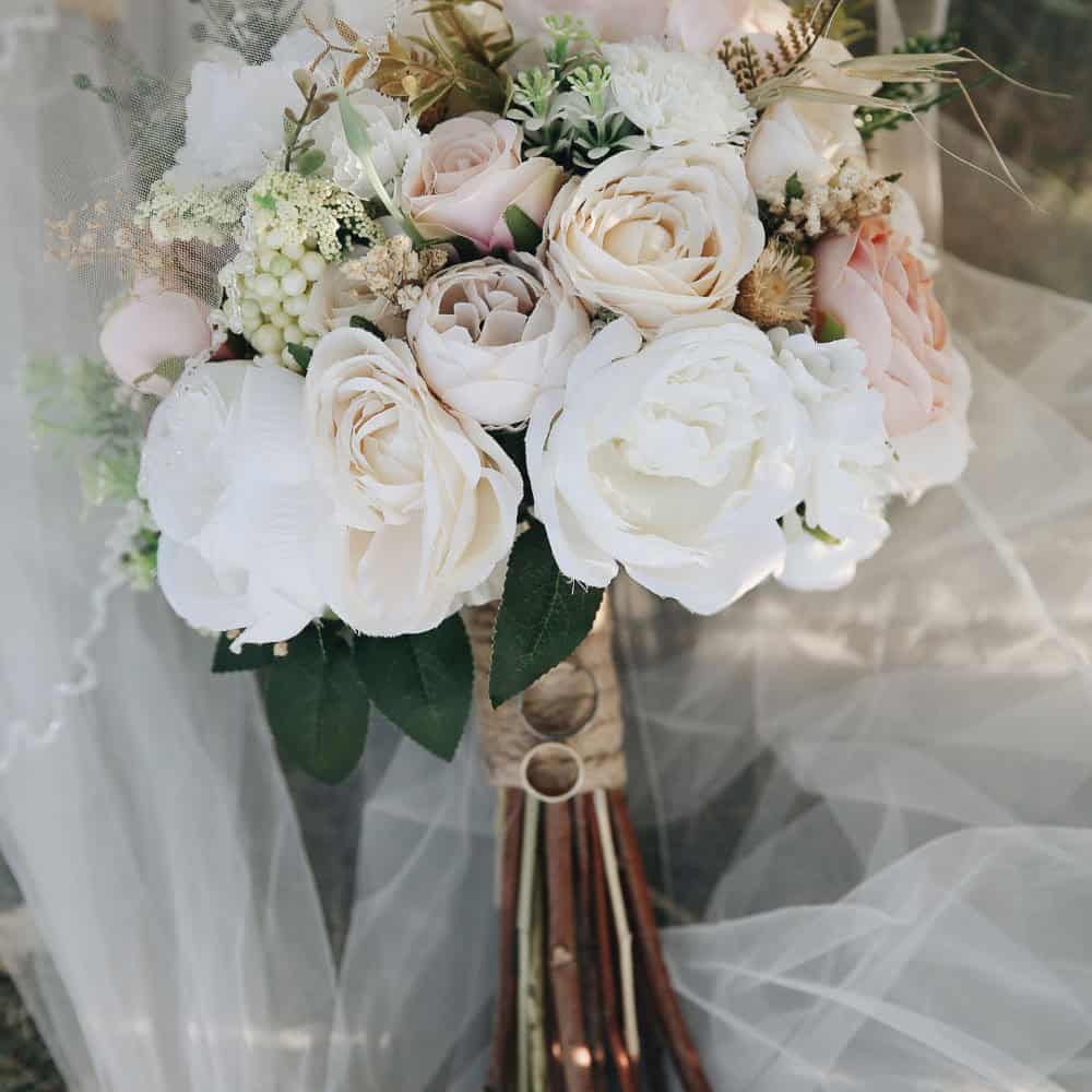 A bridal bouquet featuring white and pale pink roses, greenery, and wrapped stems, placed on white tulle fabric.