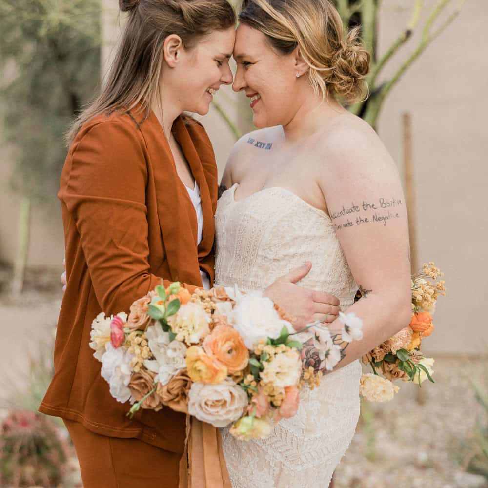 Two women embrace outdoors, smiling with foreheads touching. One wears a brown suit, the other a strapless white dress. Both hold bouquets of flowers.