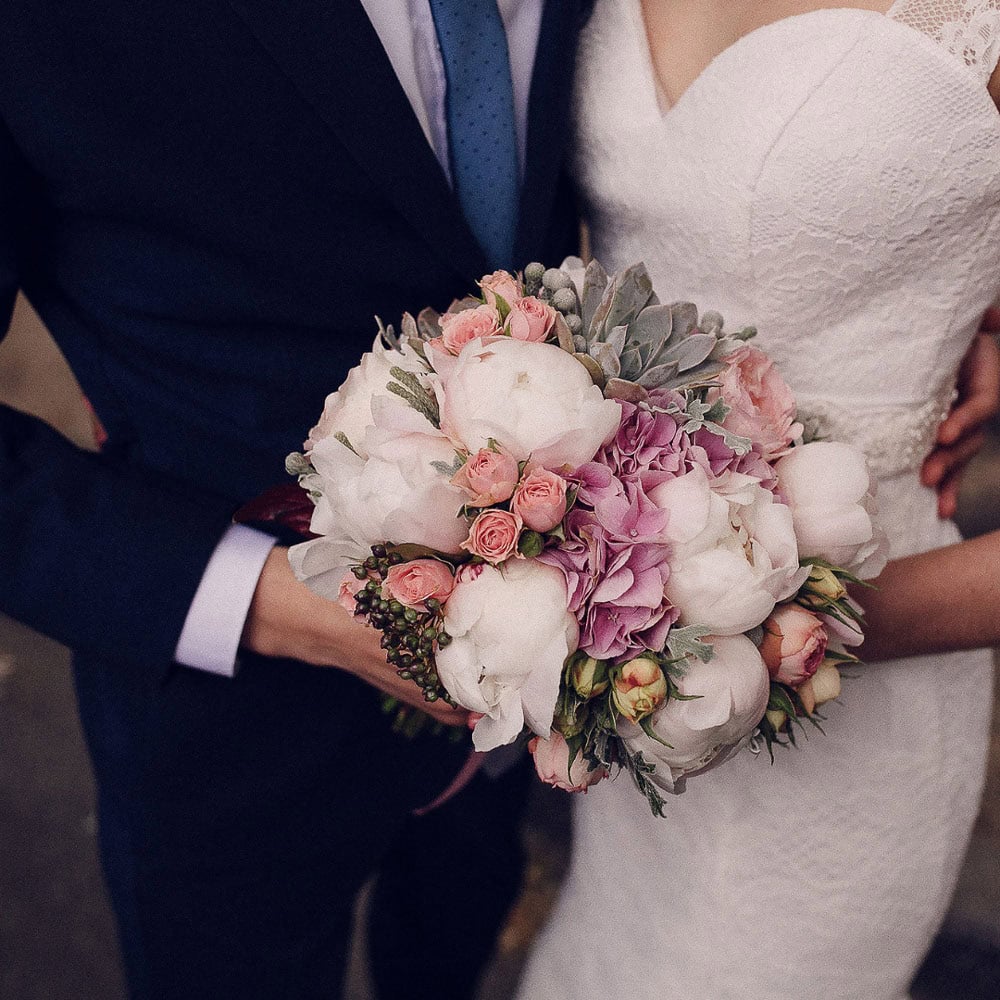 A bride in a white dress and a groom in a dark suit hold a bouquet of white, pink, and purple flowers.