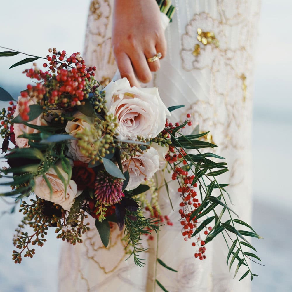 A woman in a white and gold dress holds a bouquet of pale pink roses, red berries, and greenery.
