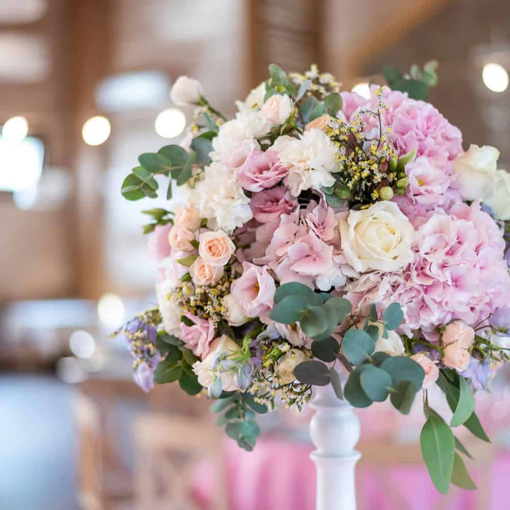A bouquet of pink, white, and cream flowers with green leaves arranged in a tall white vase, set indoors with soft lighting in the background.