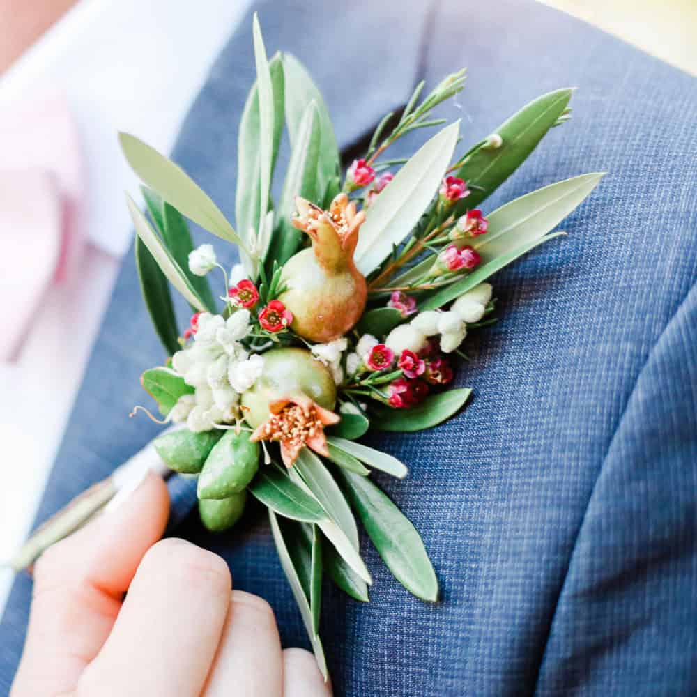 A close-up of a boutonniere with green leaves, small pomegranates, and red and white flowers pinned to a blue suit jacket.