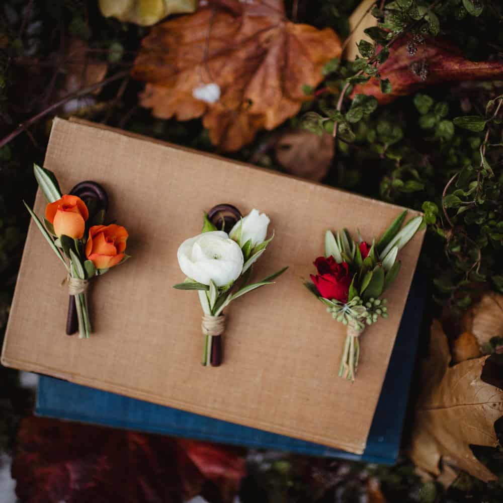 Three small boutonnieres with orange, white, and red flowers are placed on a closed book, surrounded by autumn leaves and greenery.