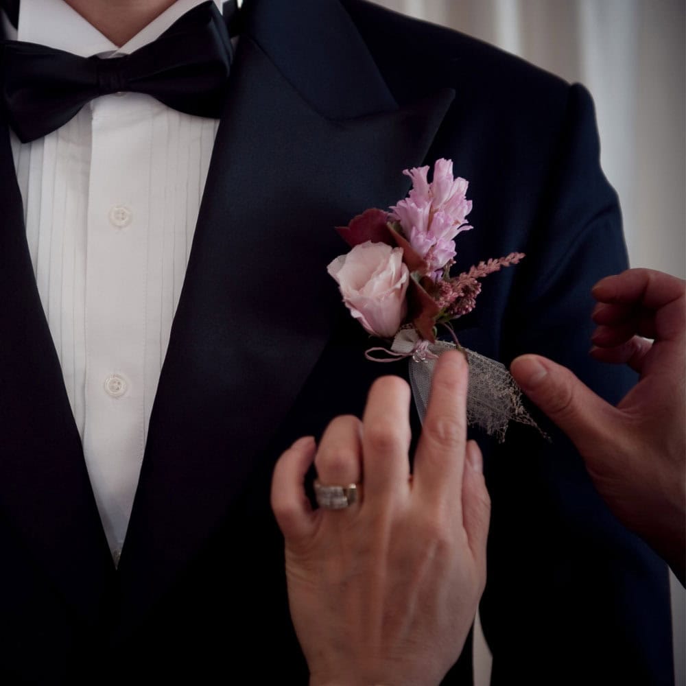 A man in a tuxedo has a boutonniere with pink flowers being pinned to his lapel by another man.