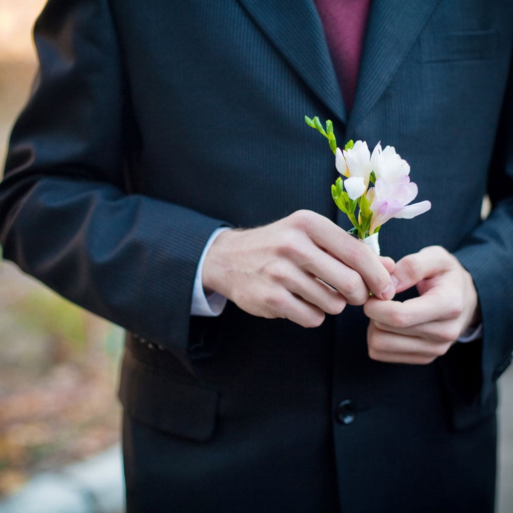 A man in a dark suit holds a small bouquet of light pink flowers, with the upper body and hands visible.
