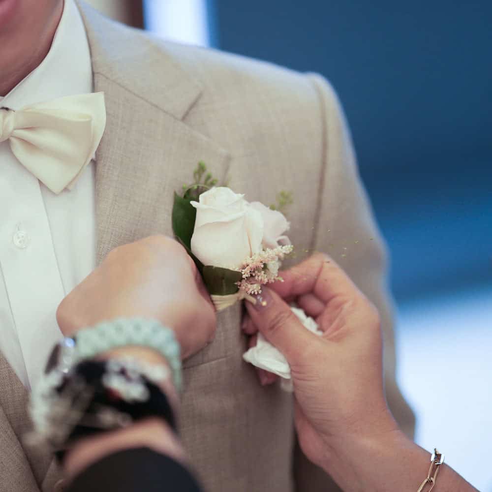 A woman pins a boutonniere with white flowers onto the lapel of someone wearing a light beige suit and a cream bow tie.