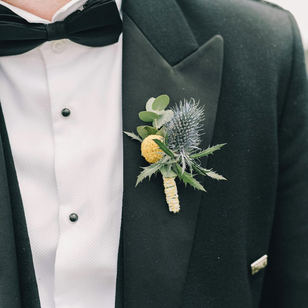 A close-up of a man in a black tuxedo with a white shirt and black bow tie, featuring a boutonniere with thistle, greenery, and a yellow billy ball.