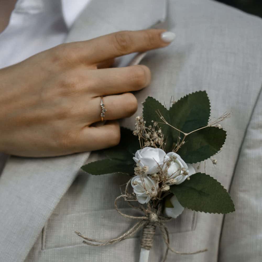 A man wearing a light suit jacket with a white floral boutonniere and holding the lapel, a ring is visible on a woman's finger.