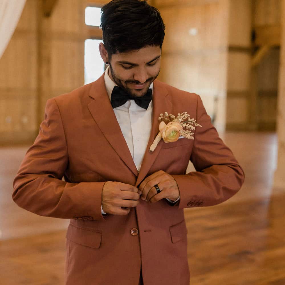 A man in a rust-colored suit with a boutonniere buttons his jacket in a wooden venue.