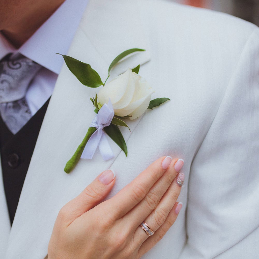 A hand with a wedding ring rests on a man wearing a white suit jacket with a white rose boutonniere pinned to the lapel.