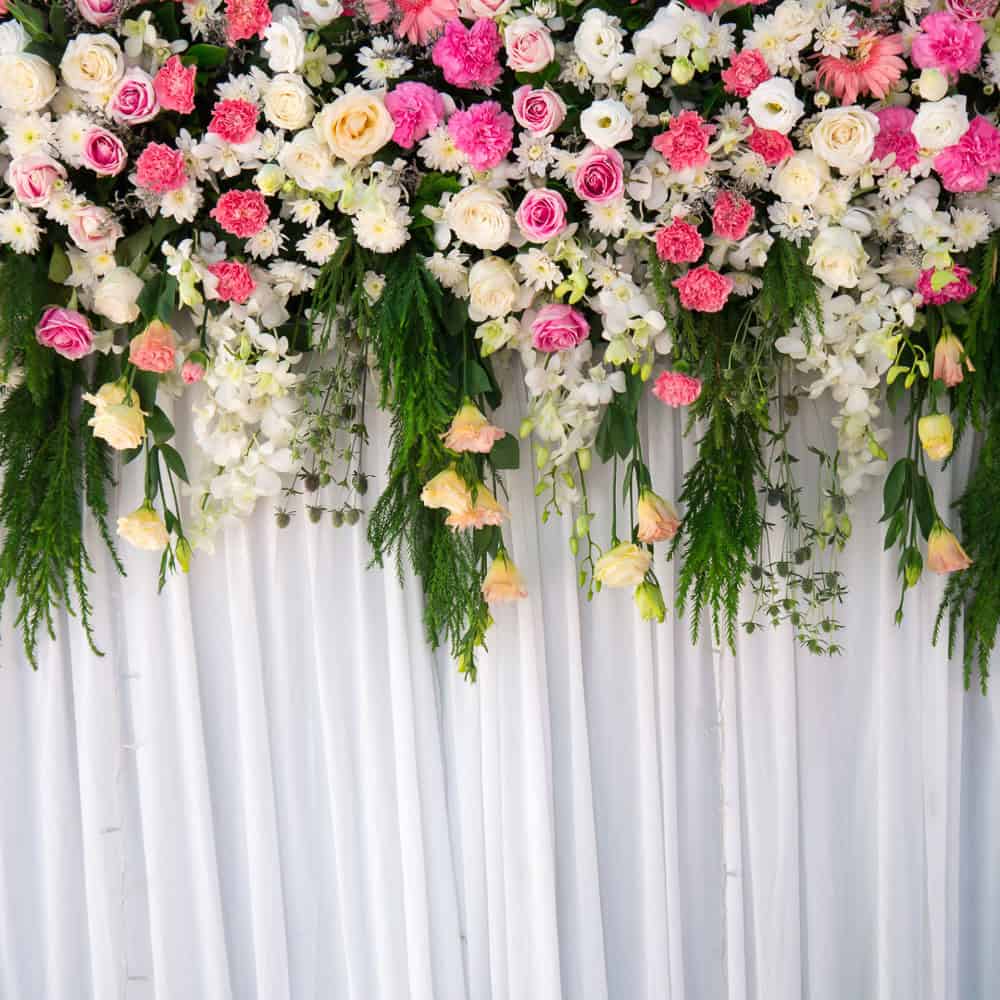 A floral arrangement of pink, white, and cream flowers with green foliage hangs above a white draped fabric backdrop.