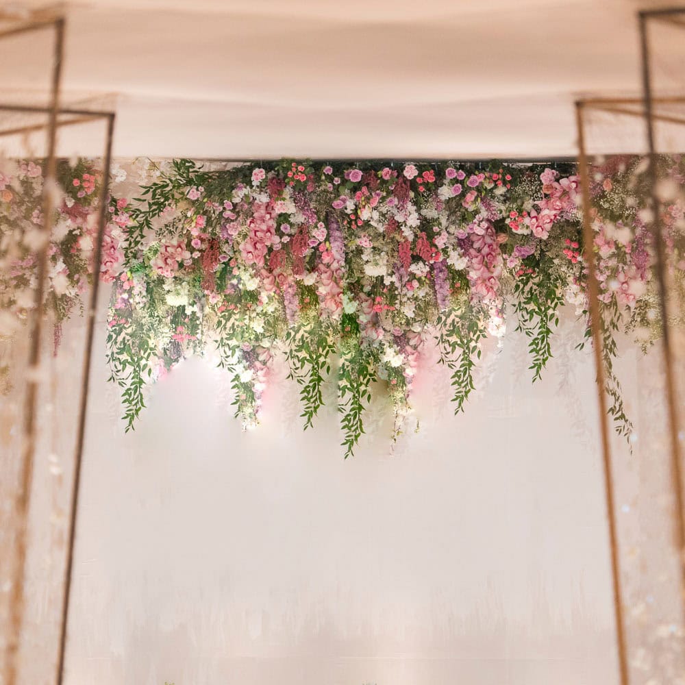 Hanging floral arrangement with pink, purple, and white flowers, along with green foliage, suspended from the ceiling against a neutral background.