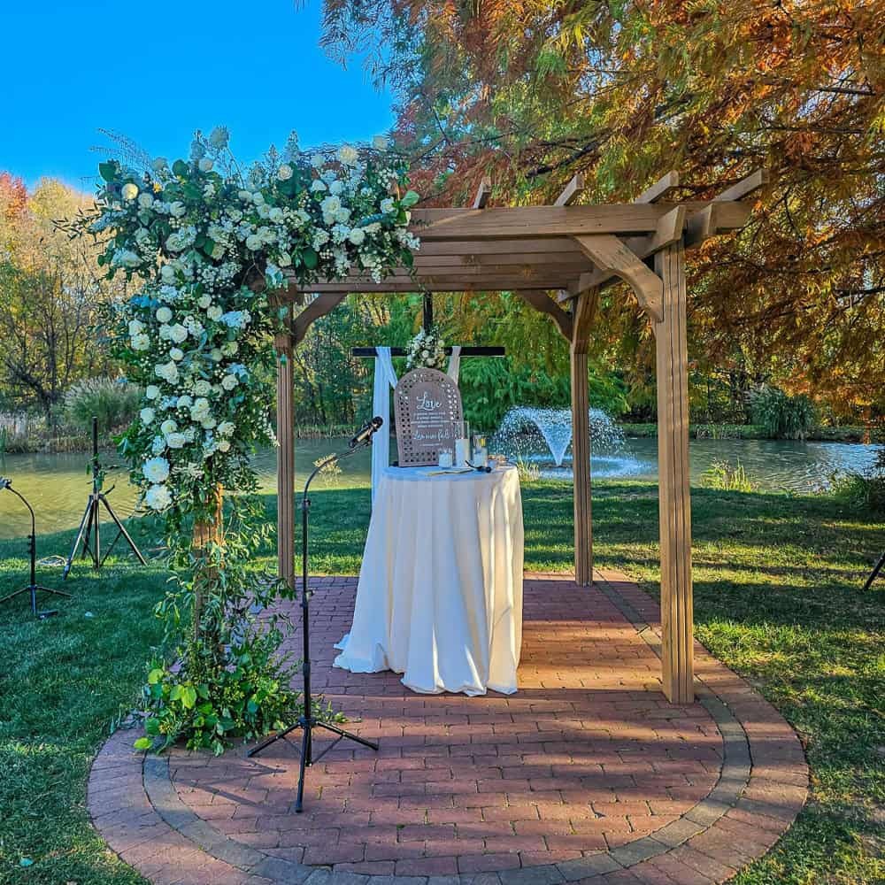 Wooden pergola decorated with white flowers and greenery stands on a brick circle near a pond with a fountain; a table and microphone are set up under it.