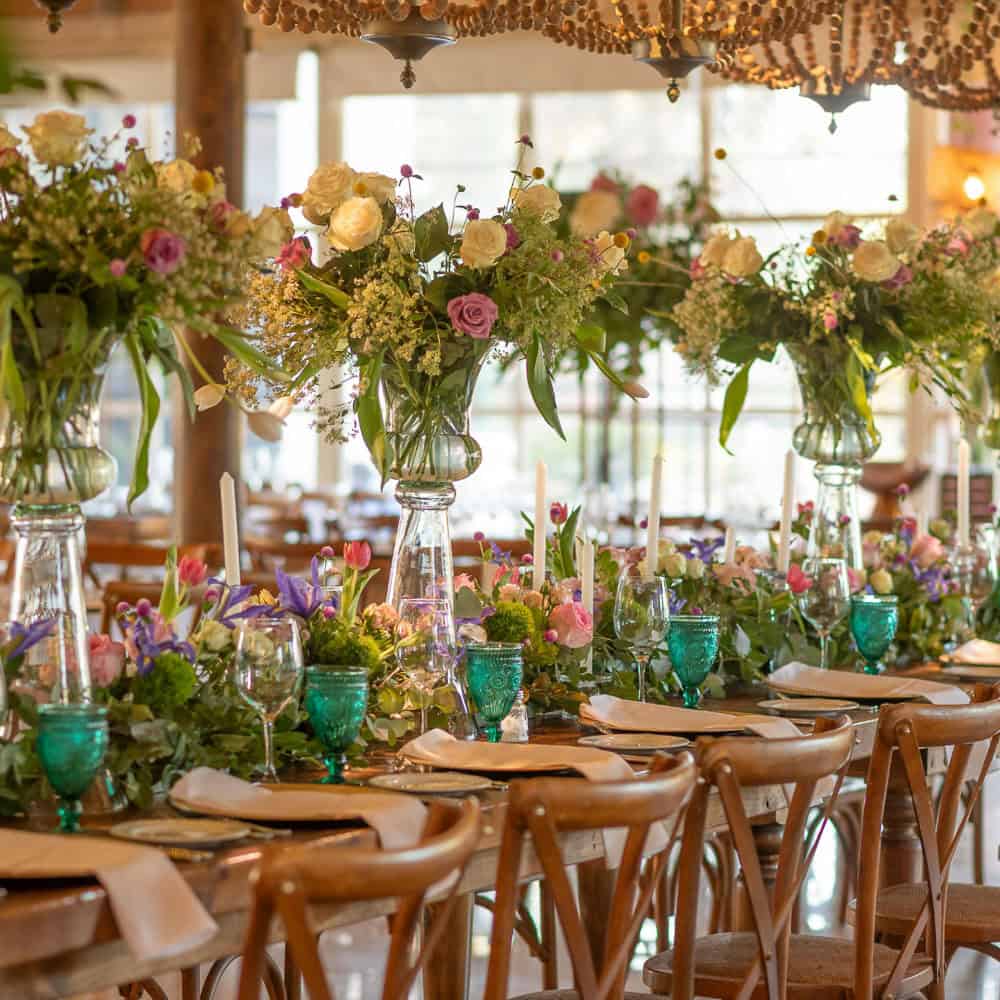 Long wood table set for an event with wooden chairs, green glass goblets, neutral plates, and large floral centerpieces with assorted flowers and greenery.