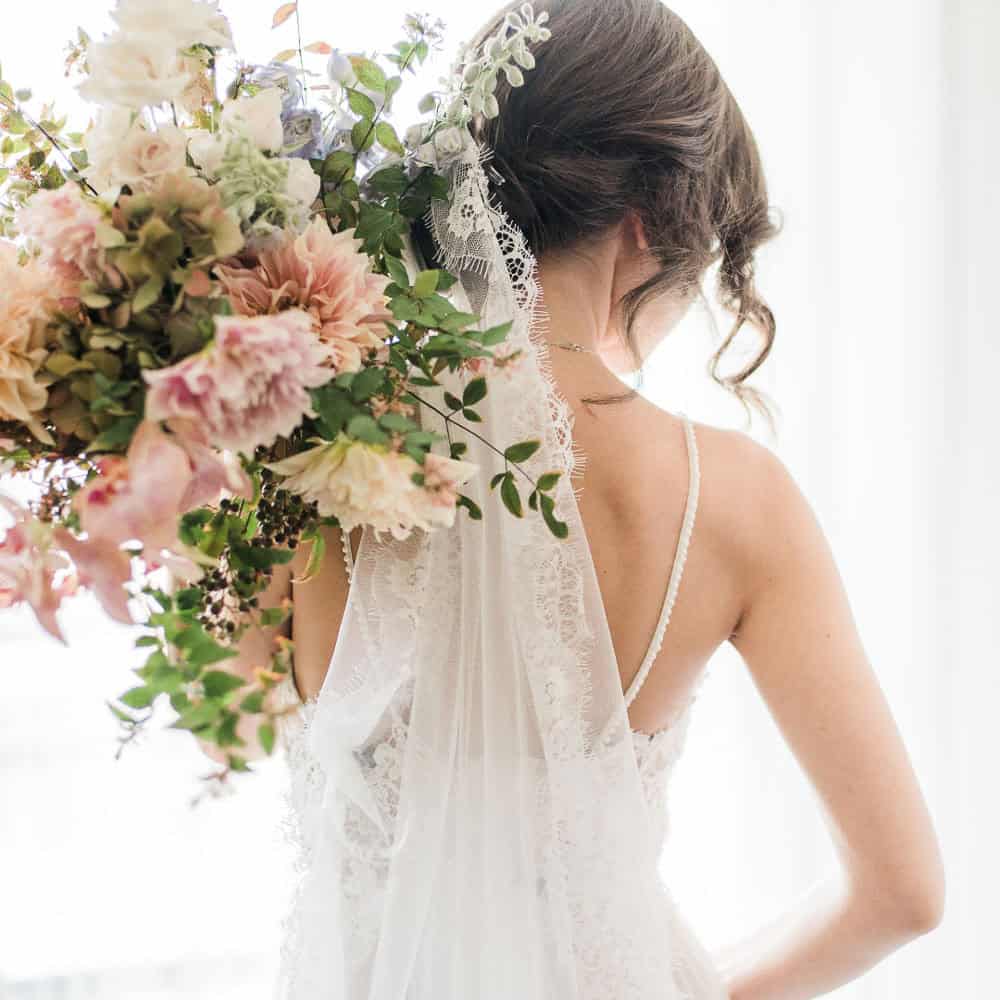 Bride wearing a lace wedding dress and veil, holding a large bouquet of pastel flowers, facing away from the camera.