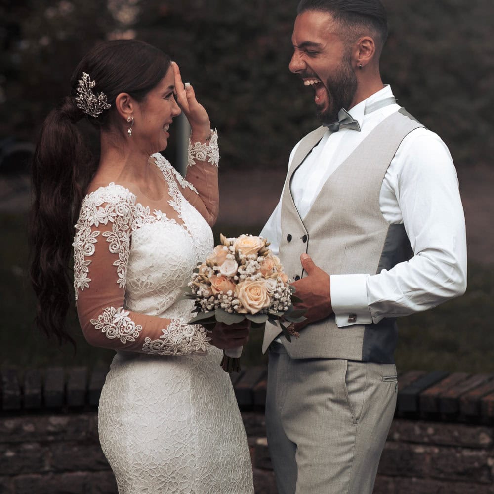 A bride in a lace wedding dress holds a bouquet and smiles while a groom in a light suit vest laughs; they face each other outdoors.